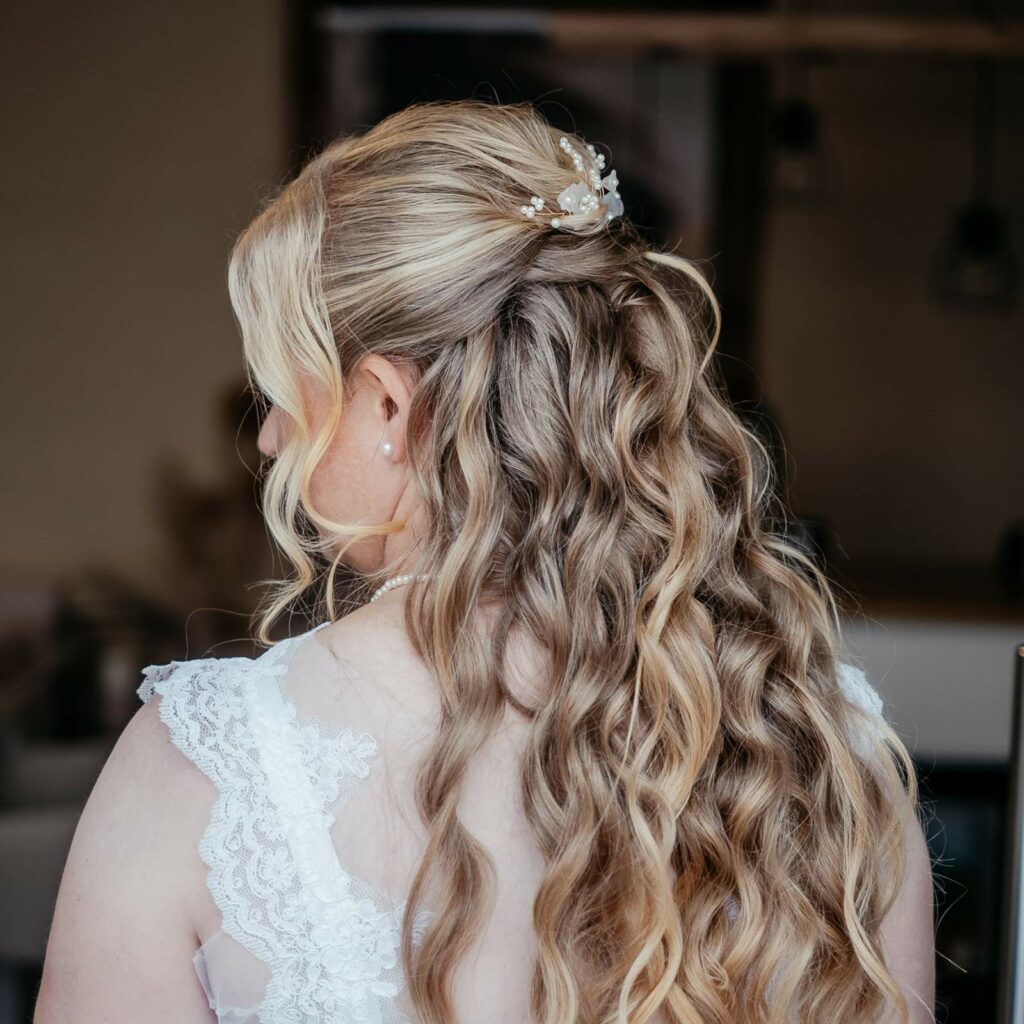 Das Bild ist ein Beispiel für eine halboffene Brautfrisur mit Beach Waves in blonden Haaren und wunderschönem Haarschmuck.
