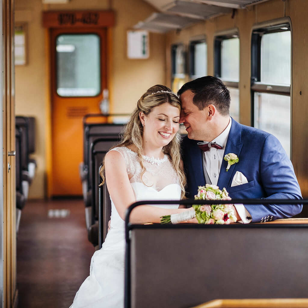 Das glücklich lächelnde Brautpaar sitzt in einem Waggon der Weißeritztalbahn am Bahnhof Dippoldiswalde. Die Braut trägt ein dezentes Braut-MakeUp in natürlichen Farben.