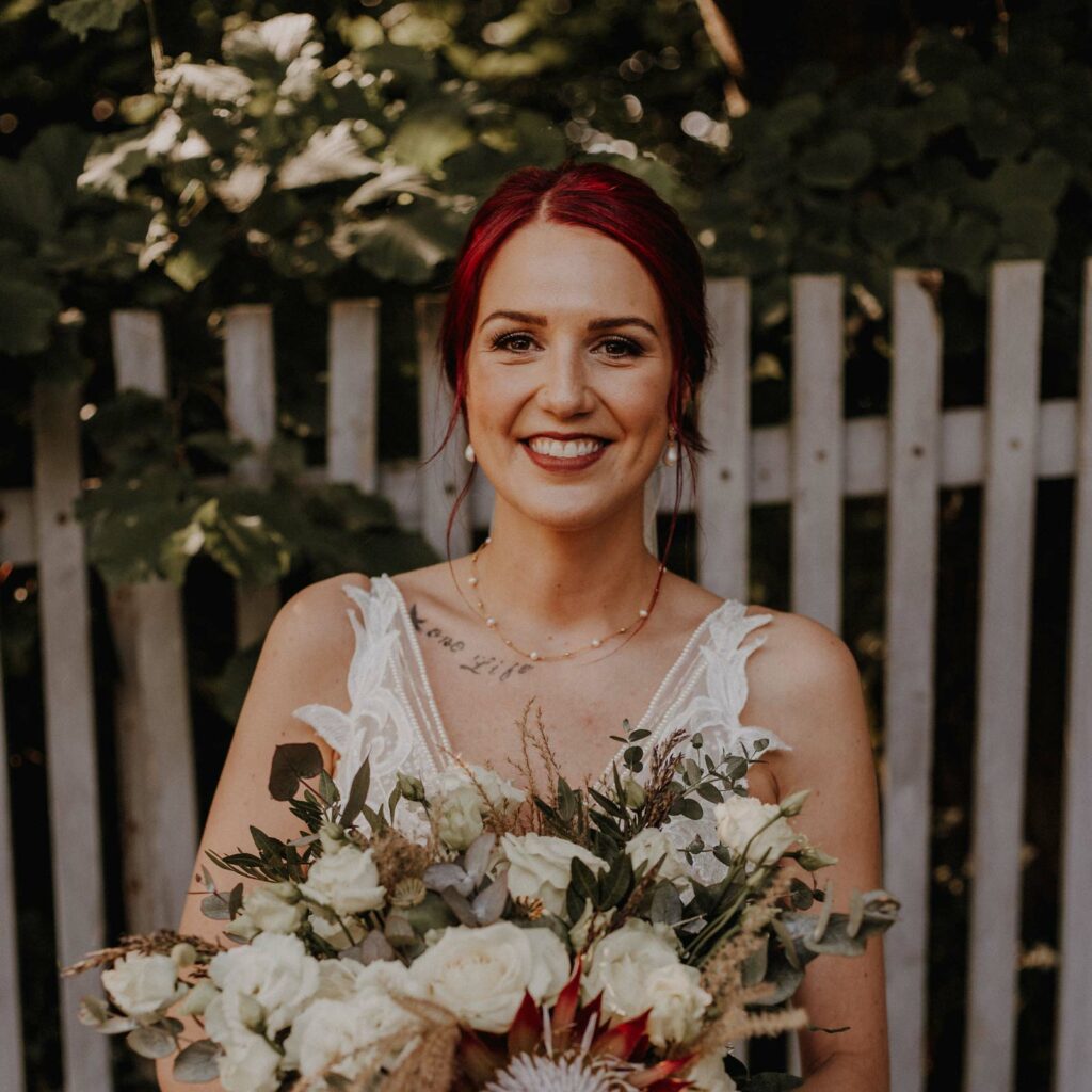 Eine glücklich strahlende Braut steht vor einem Holzzaun. Die Braut trägt ein ausdrucksstarkes Braut-MakeUp mit einem kräftigen roten Lippenstift, der perfekt zu den roten Haaren passt.