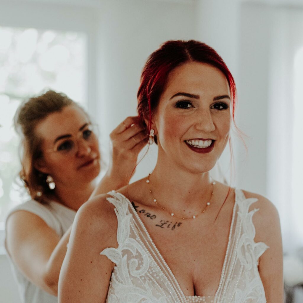 Eine wunderschöne Braut lächelt glücklich am Morgen ihres Hochzeitstages, während ihre Haare gemacht werden. Sie trägt ein ausdrucksstarkes Braut-MakeUp mit einem kräftigen roten Lippenstift, der perfekt zu den roten Haaren passt.