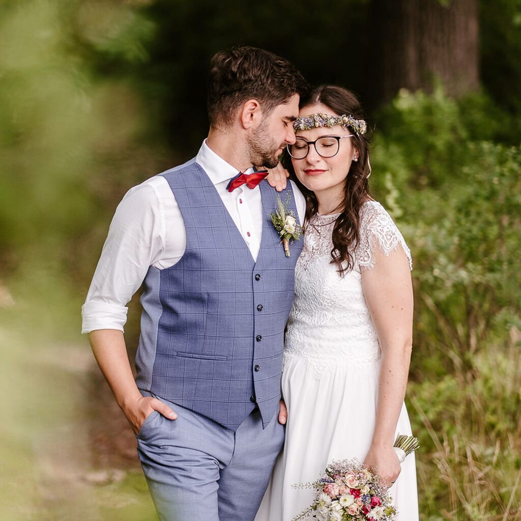 Ein wunderschönes Brautpaar im Grünen. Die Braut trägt leichte Wellen und einen Blumenkranz im Haar. Das klassische Braut-MakeUp wird von einem kräftigen rotenLippenstift abgerundet.