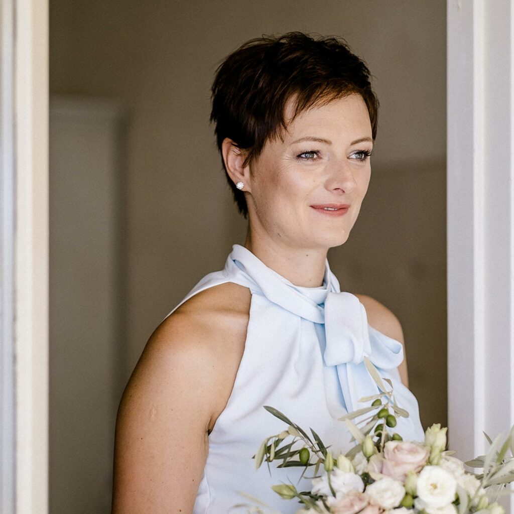 Eine Braut mit kurzen Haaren und typgerechtem, dezentem Braut-MakeUp steht lächelnd am offenen Balkonfenster. Sie hält einen Brautstrauß in zarten Pastelltönen in der Hand.