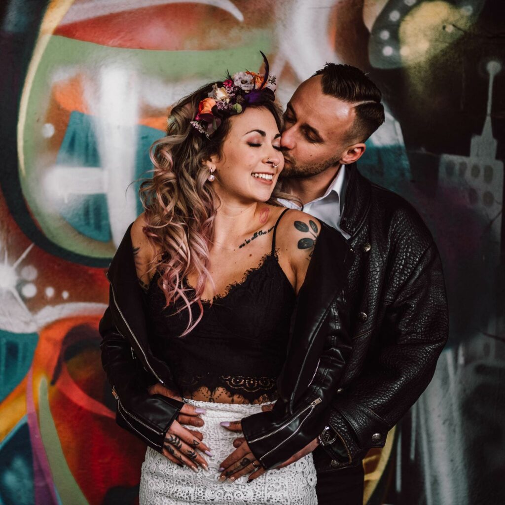 Brautpaar in Lederjacken vor einer Graffiti-Wand. Die Braut trägt die Haare in offenen Locken mit einem farbenfrohen Blumenkranz. Sie ist ausdrucksstark geschminkt.