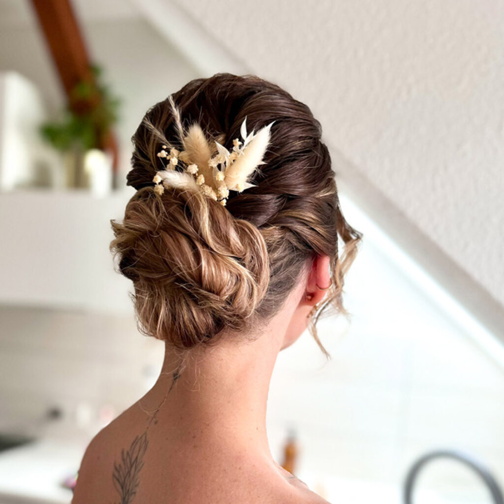 Ein tiefer Dutt an dunkelblondem Haar mit gelocktem Hinterkopf und Haarschmuck aus Trockenblumen.